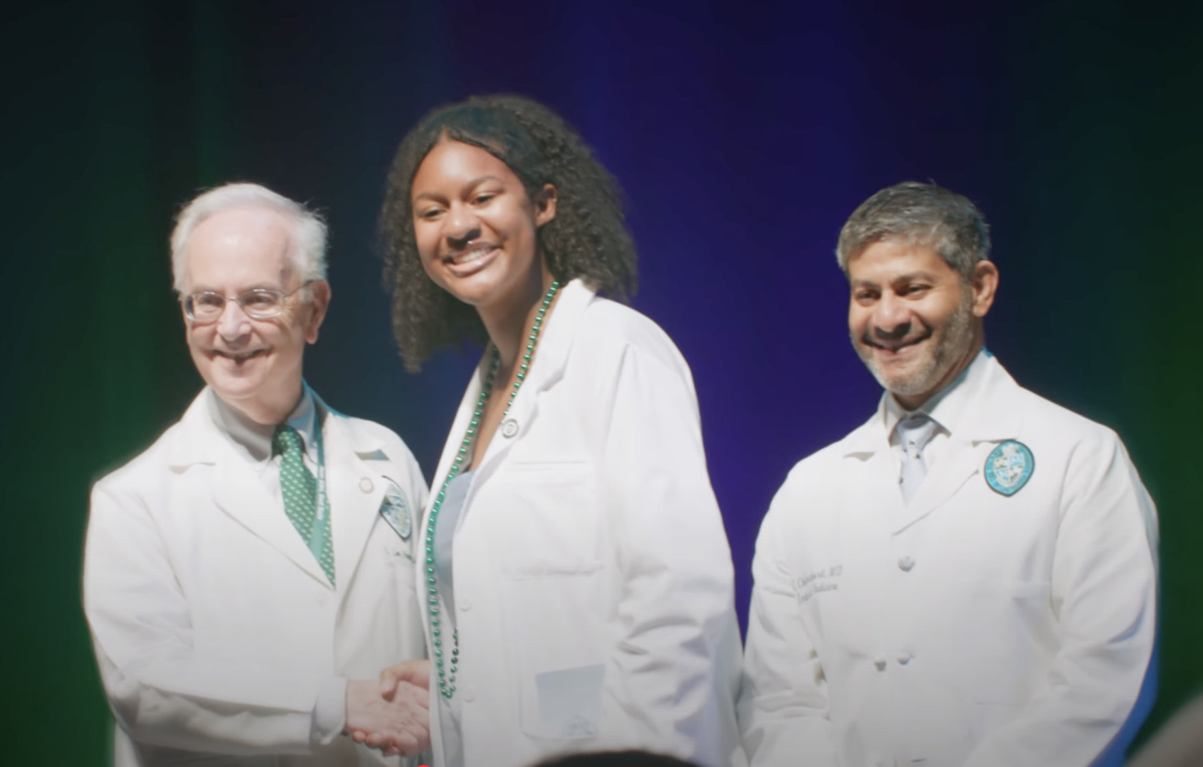 A woman in a short white doctors coat shakes the hand of a man in a doctors coat while another man in a doctors coat smiles