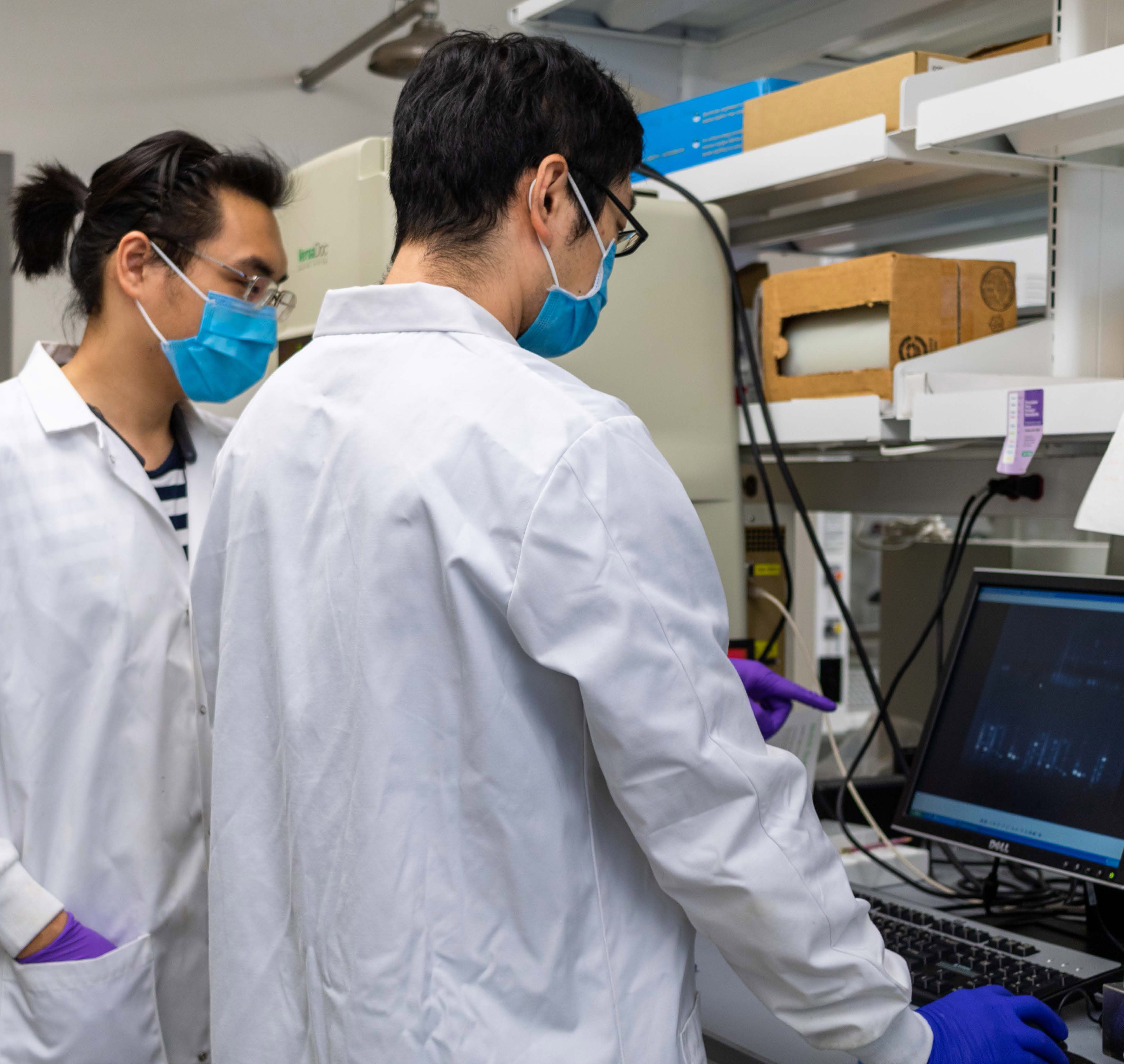 two men in lab coats review research on a computer screen