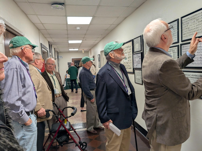 Men wearing green baseball caps and sport coats stand in a hallway looking at composite photos on the wall