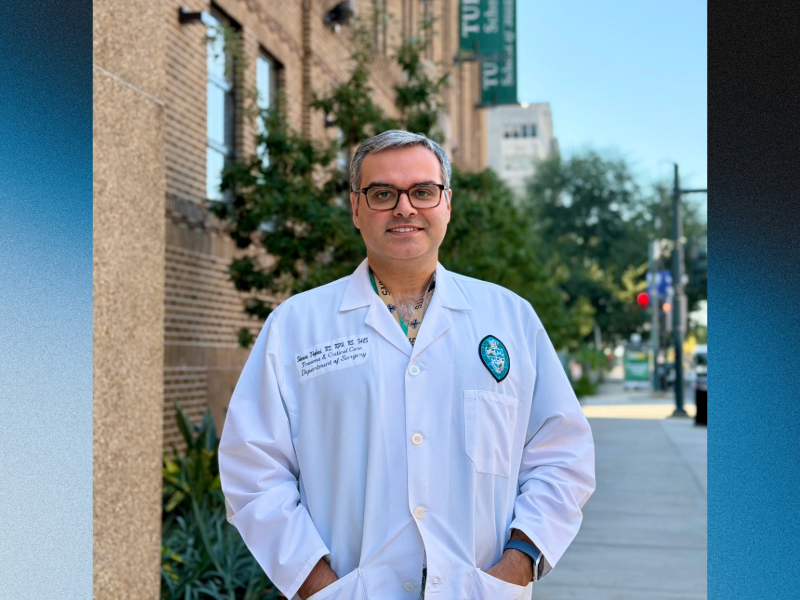 A photo of Sharven Taghavi standing outside of a beige brick building wearing a white doctor's coat