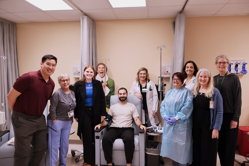 Group of 10 smiling people, including medical staff, surrounding a man in a clinic chair.