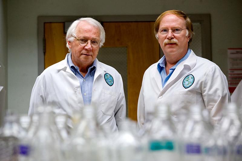 two men in lab coats stand behind glass flasks