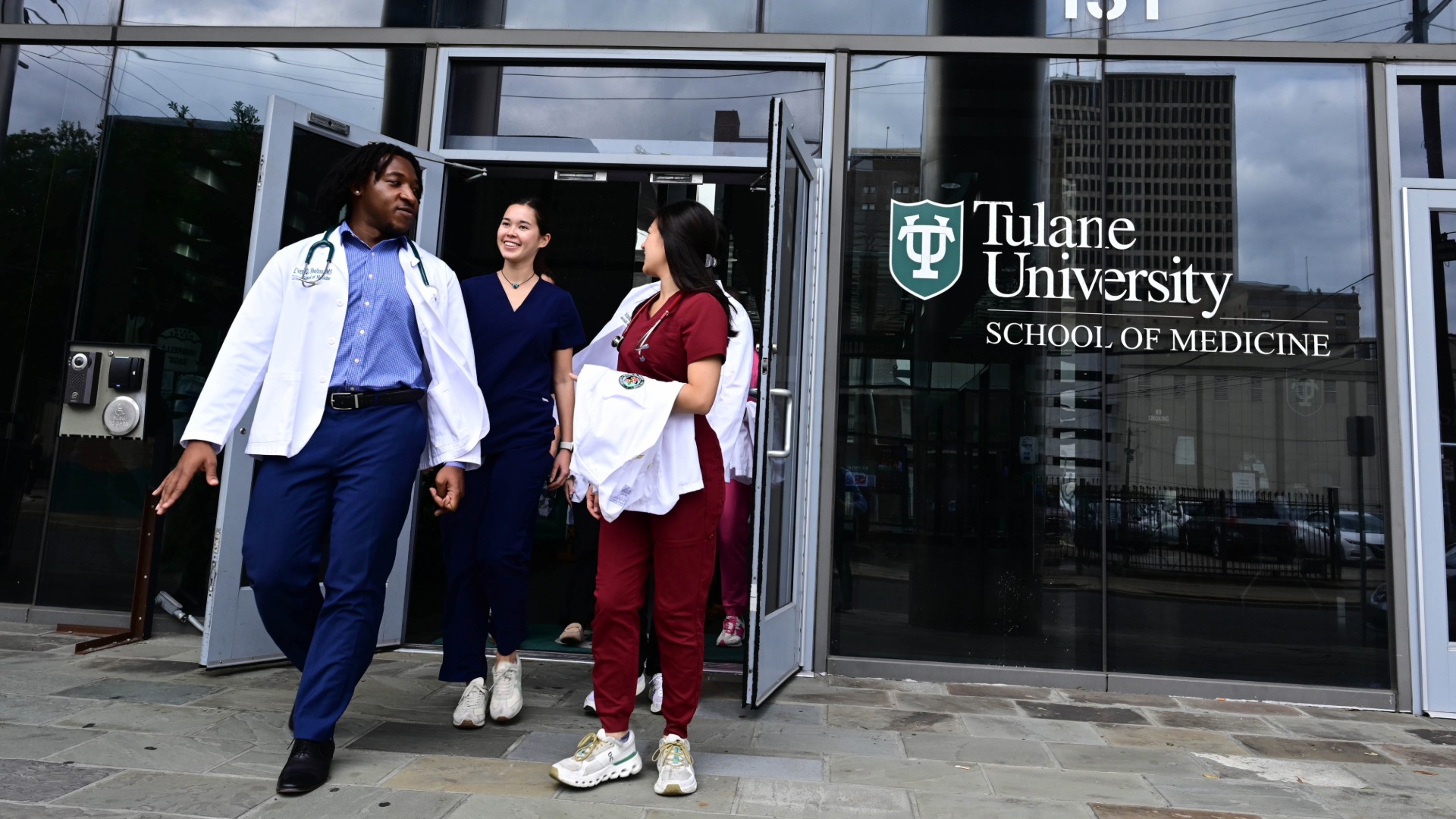 Three students walk out of building, One student is wearing a white doctors coat