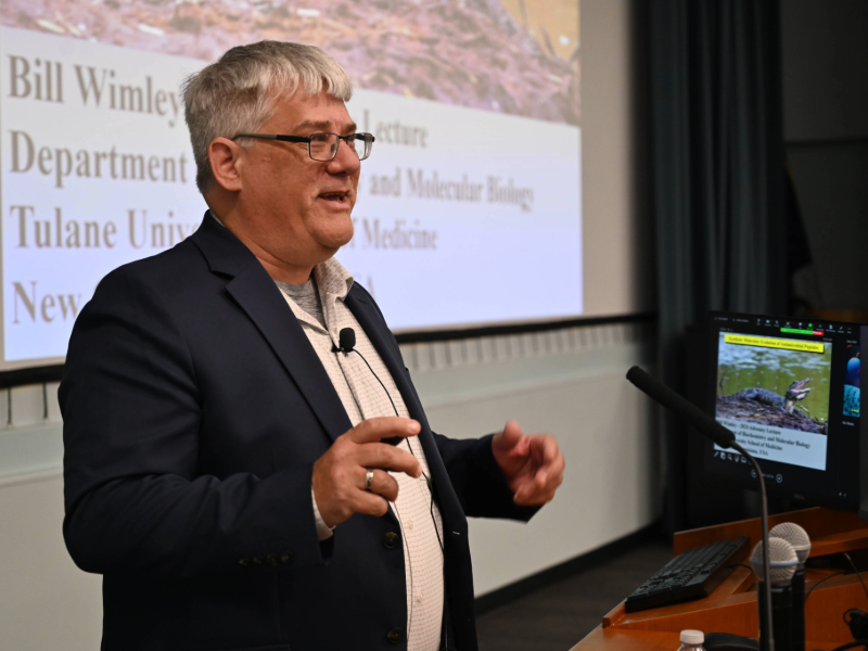 A man wearing a black coat and white shirt stands at a podium with a screen behind him
