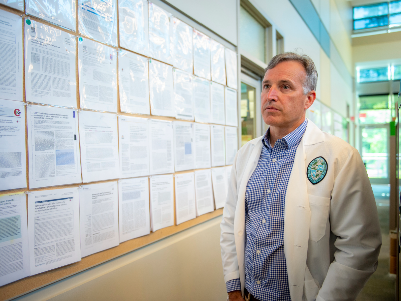 A man in a white lab coat looks over a bulletin board covered in research papers