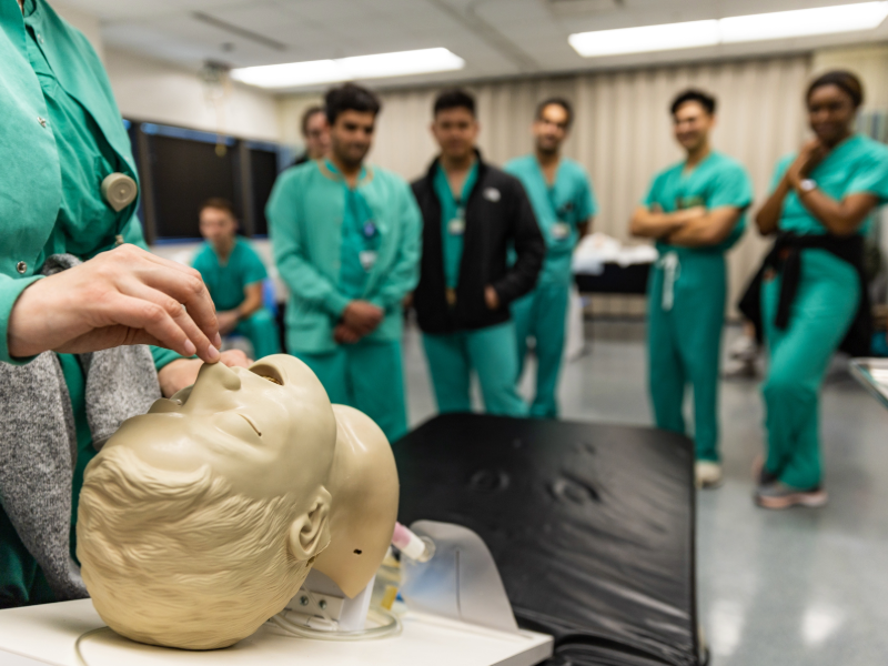 Residents in green scrubs watch as a healthcare worker inserts a breathing tube into a simulator