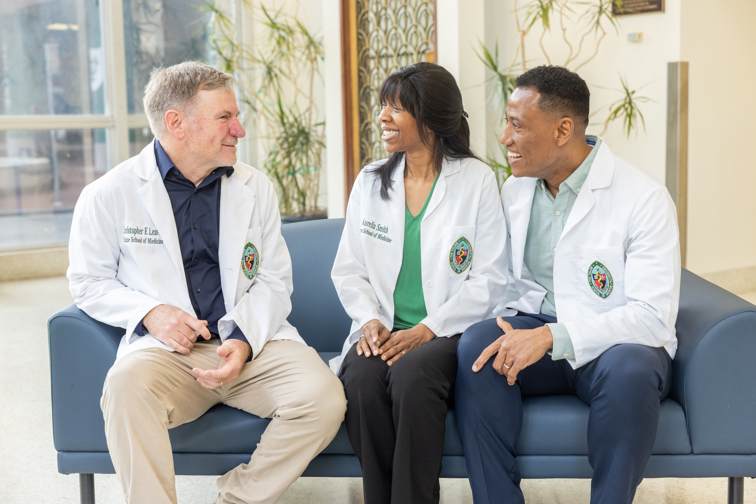 Three medical students wearing short white lab coats sit on a couch talking to each other
