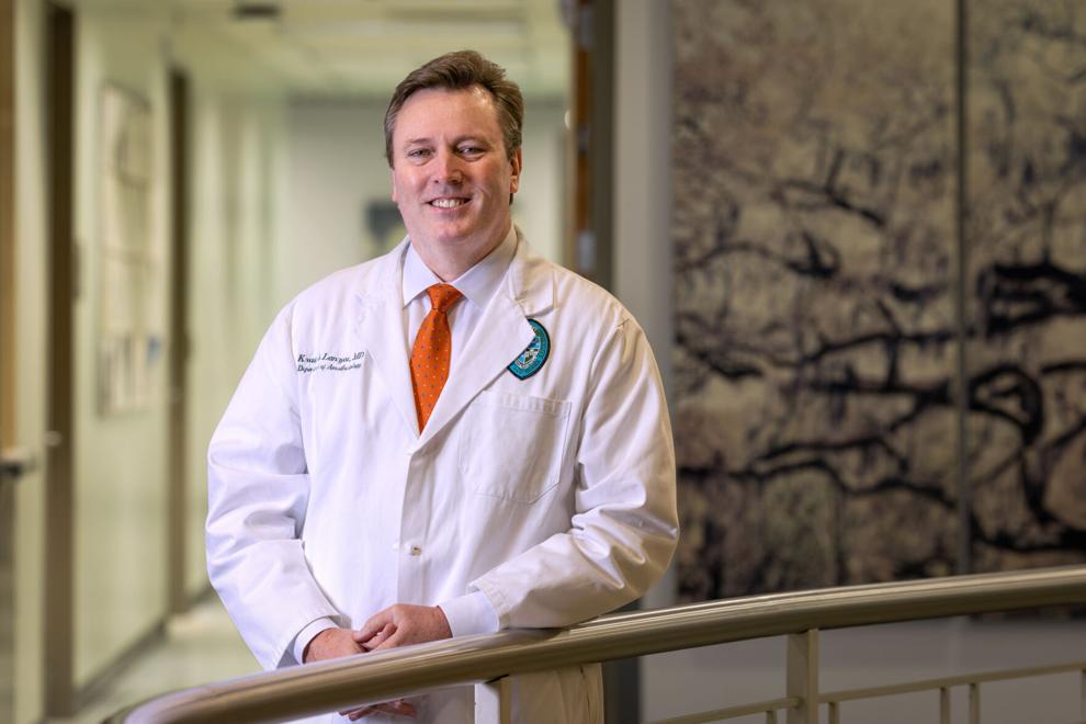 a man stands against a railing smiling and wearing a doctors coat