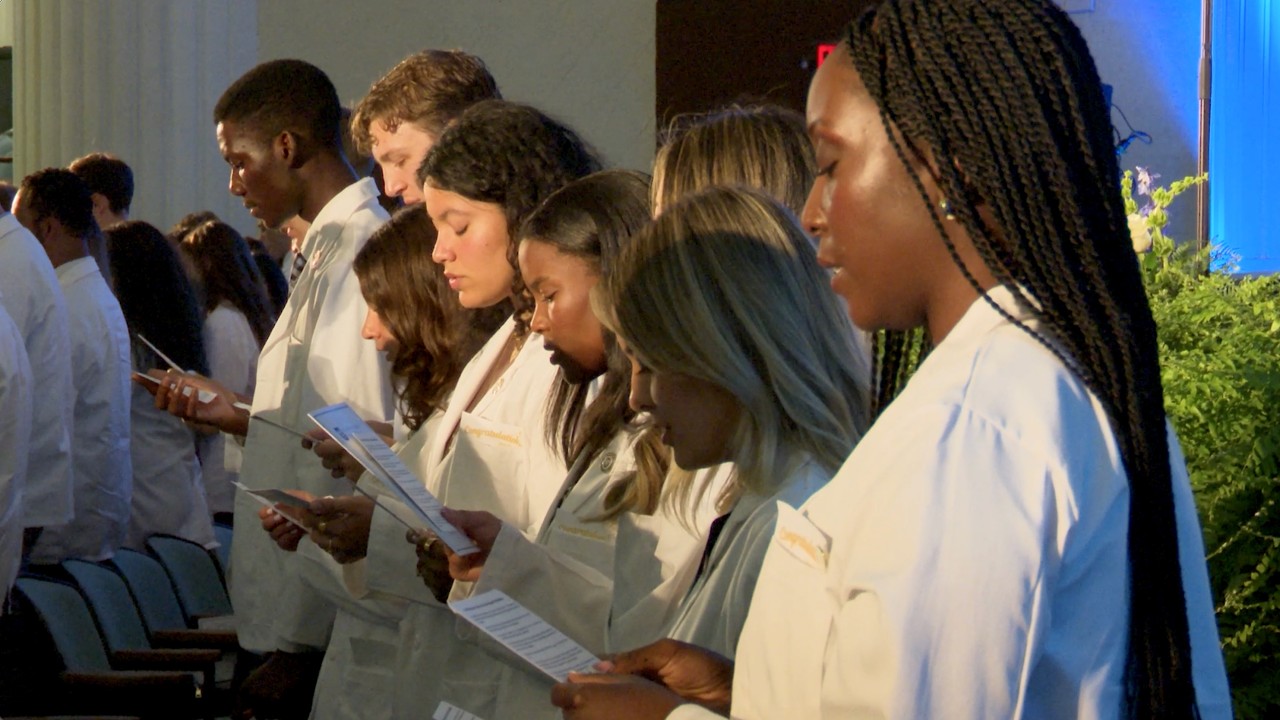Photograph of students in white coats reading papers during a ceremony.