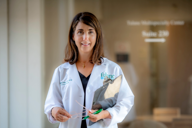 Smiling female doctor in a white lab coat holding a clipboard and glasses.