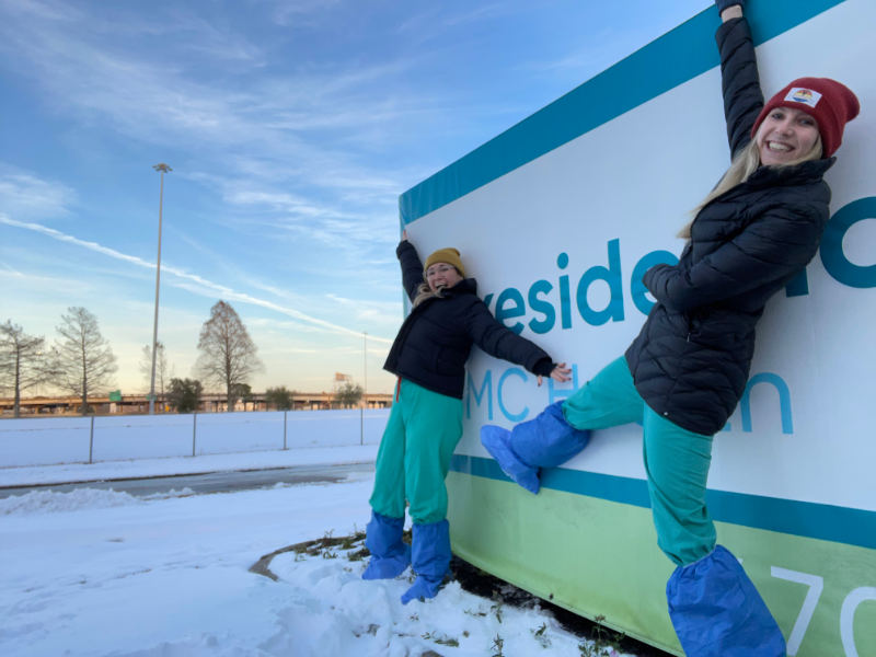 Two people in medical scrubs pose by a sign in the snow