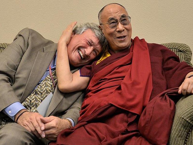 A smiling man in a suit is hugged by the Dalai Lama