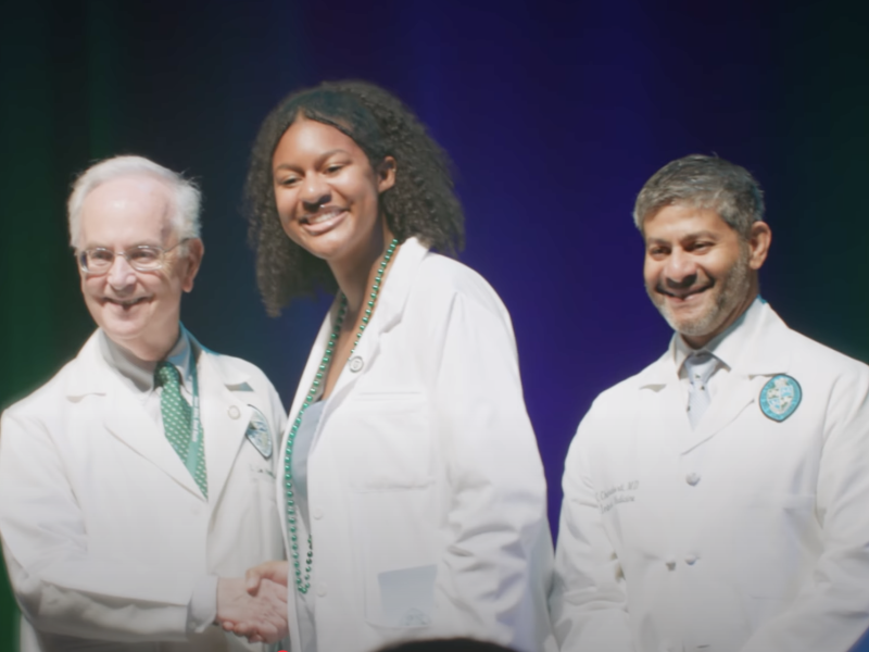A woman in a short white doctors coat shakes the hand of a man in a doctors coat while another man in a doctors coat smiles