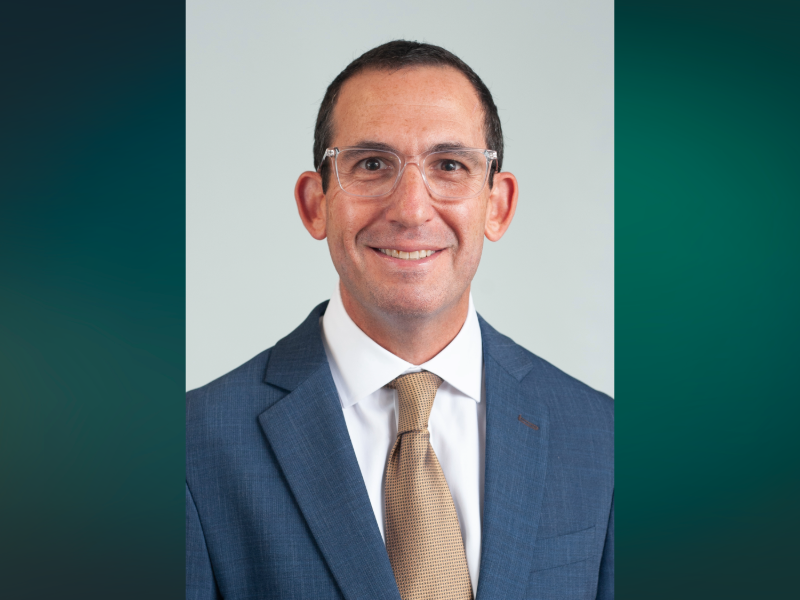 A headshot of a man in a blue suit coat with a gold tie and glasses