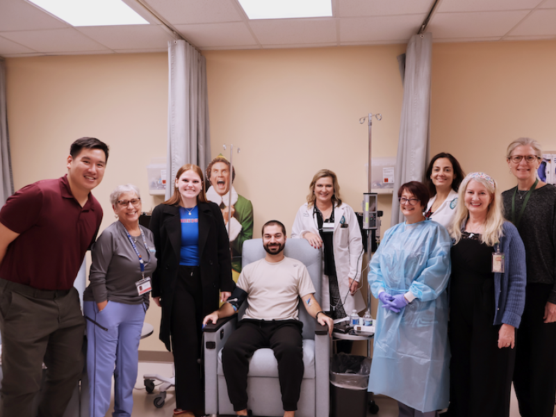 Group of 10 smiling people, including medical staff, surrounding a man in a clinic chair.