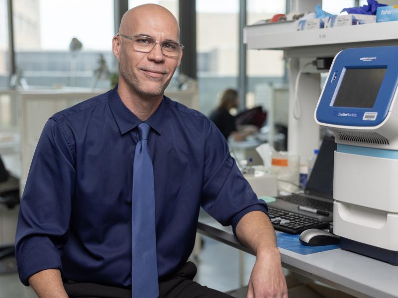 Mark Mondrinos in glasses, blue shirt, and tie smiles in a laboratory with equipment.