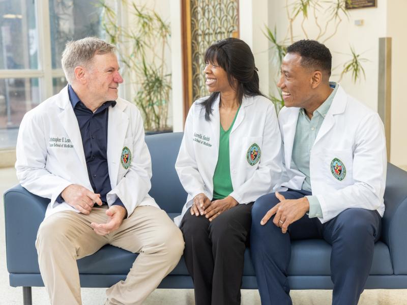 Three medical students wearing short white lab coats sit on a couch talking to each other