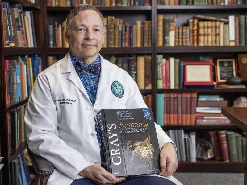 A man in a white lab coat sits in a library setting holding a copy of Gray's Anatomy