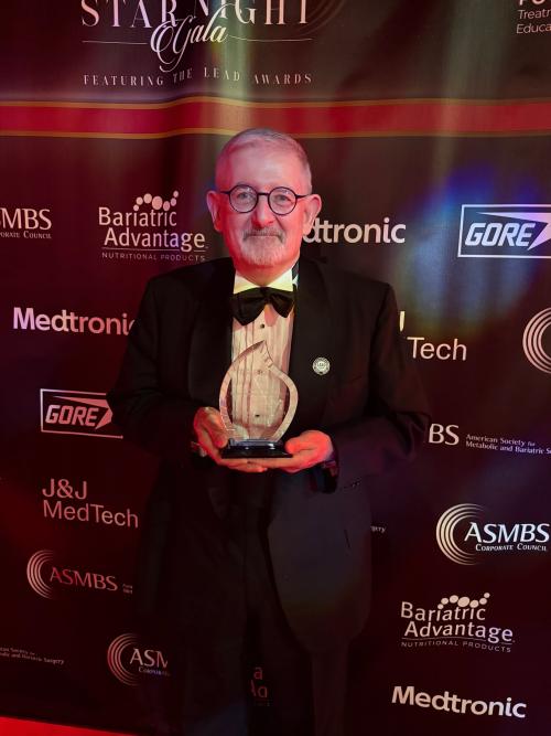 John Baker award A man in a suit and bowtie holds an award in front of a step and repeat