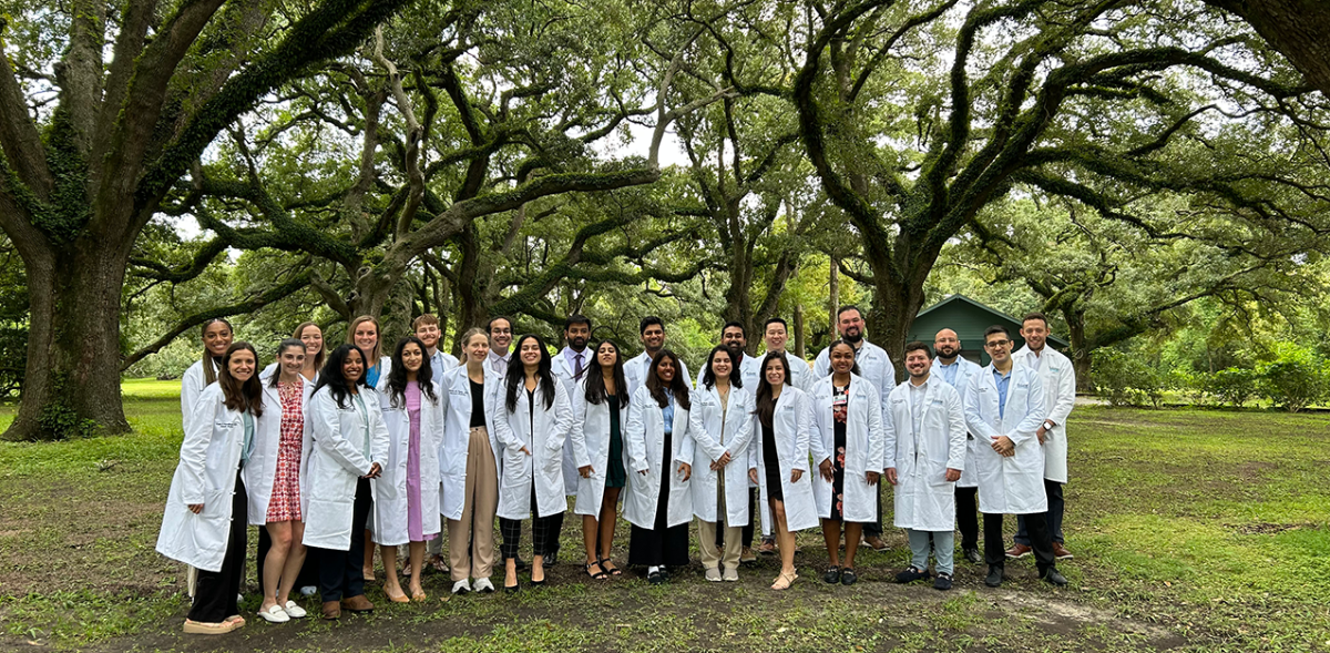 Internal Medicine Residency Banner, a group of residents in while doctor coats stand outside in a park under some green oak trees