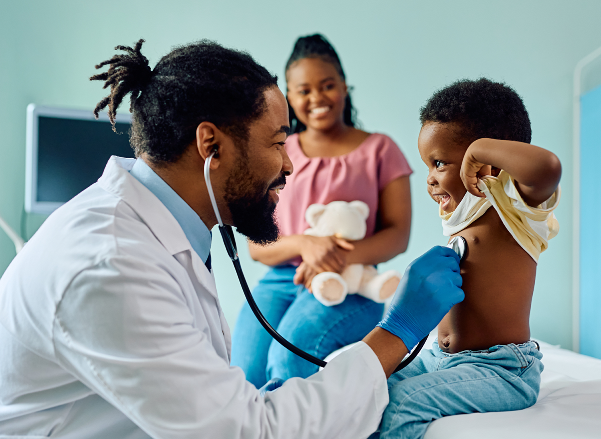 Black doctor smiles while using stethoscope on smiling young boy, mother watches proudly.