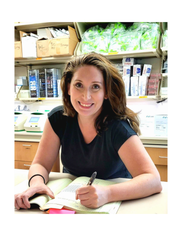 Photo depicts Catherine Weindel, PhD sitting at a lab work bench while smiling and holding a pen over a book