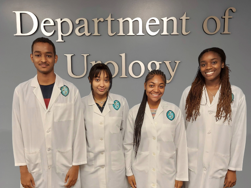 This photo shows four students standing in front of the Department of Urology sign