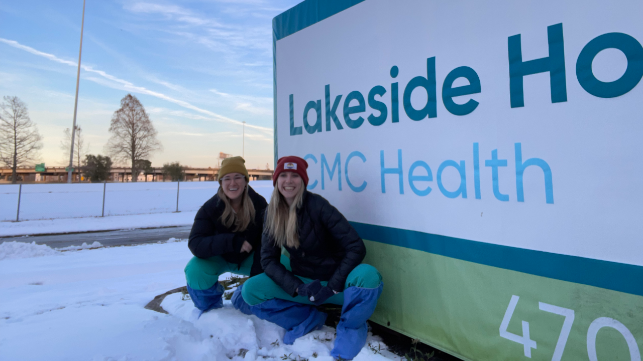 Two people in medical scrubs pose by a sign in the snow