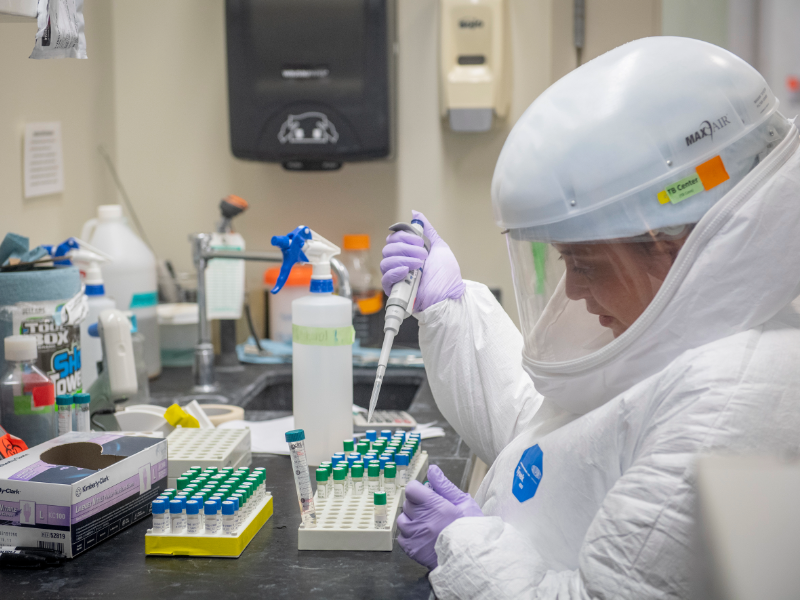 A scientist in a full hazardous materials lab suit holds a pipette over test tubes
