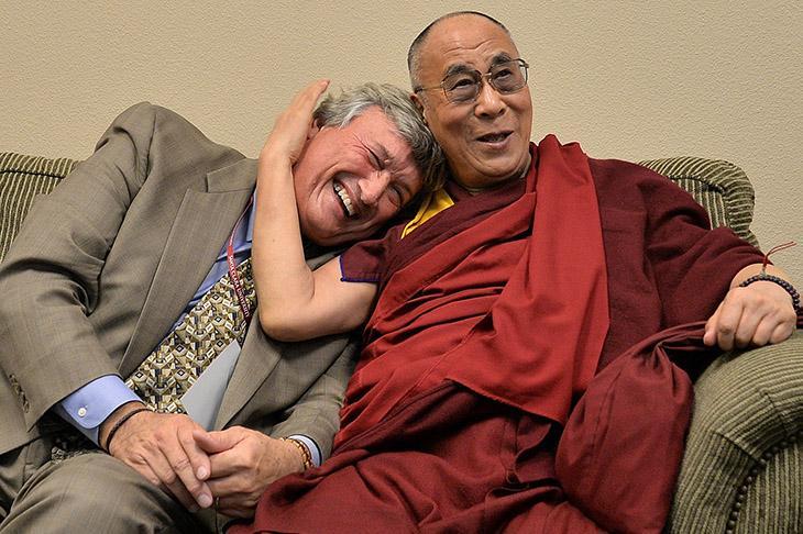 A smiling man in a suit is hugged by the Dalai Lama