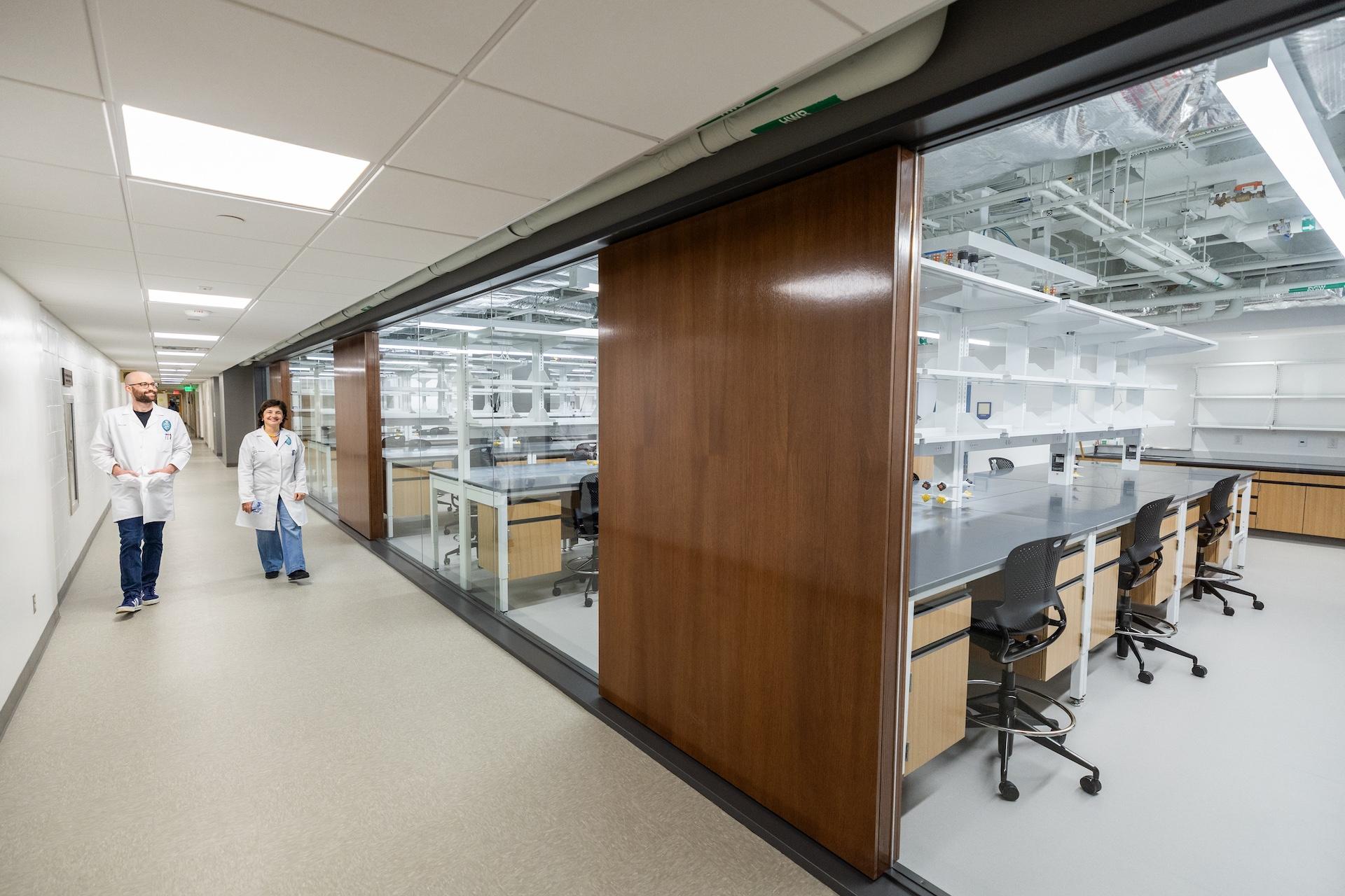 Two researchers walk down a hallway with glass-walled research labs visible behind them in a renovated academic space.