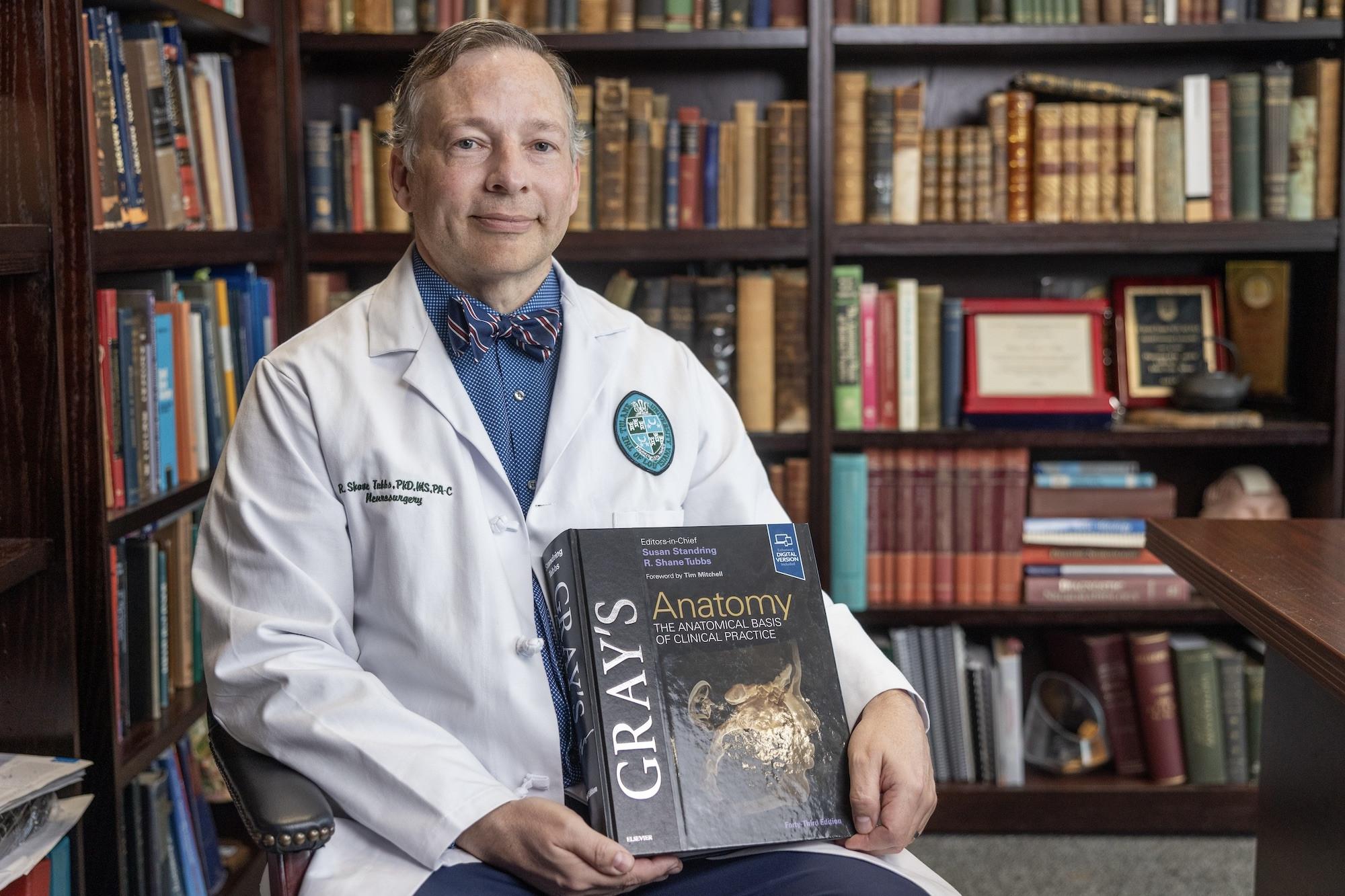 A man in a white lab coat sits in a library setting holding a copy of Gray's Anatomy