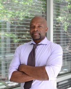 Dr. Herbert pictured with crossed arms in front of a window. He is wearing a light purple button up shirt and tie. 