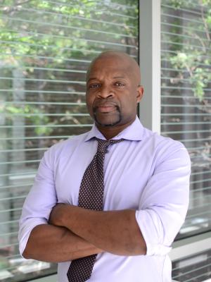Dr. Herbert pictured with crossed arms in front of a window. He is wearing a light purple button up shirt and tie. 