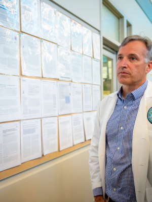 A man in a white lab coat looks over a bulletin board covered in research papers