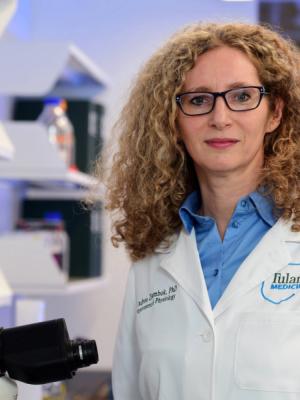 A portrait of Andrea Zsombok in a lab. She is wearing a blue shirt under a white lab coat